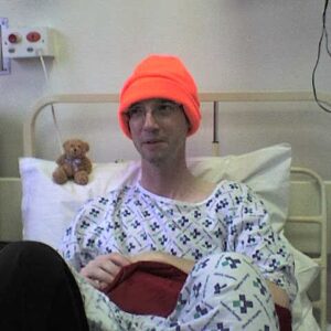 Stephen is sat in a hospital bed, wearing a fluorescent orange beanie hat, a hospital gown and holds his christmas stocking in his hand, on Christmas Day. There is a small teddy bear propped up on the top of his pillow, next to him.