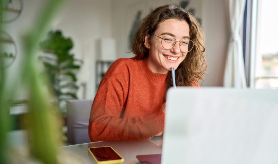 A young woman is sat at a desk looking at her laptop. She is holding a pen and her mobile phone is next to her. She is smiling.