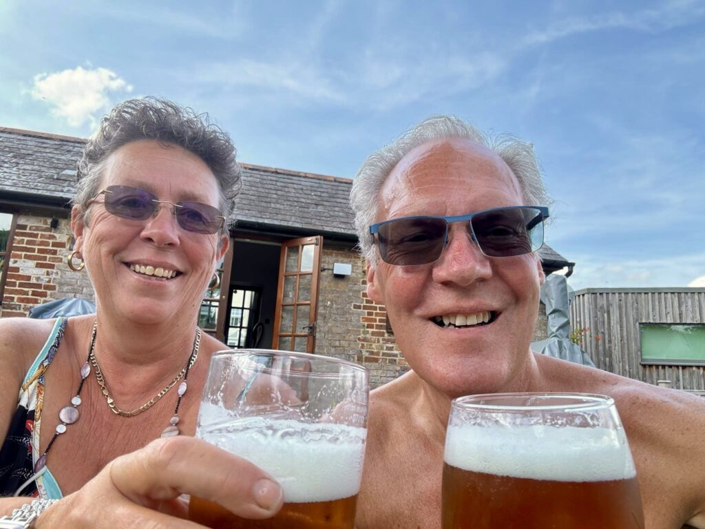 Sarah and Graham are sat enjoying a pint of beer each outside. They are both wearing sunglasses and the sky is blue.