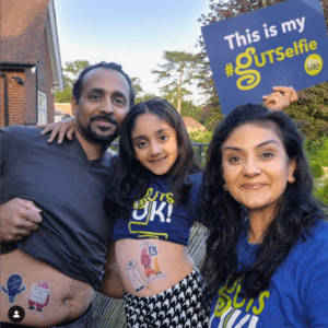 A family of three smile for the camera when taking a selfie. One parent is holding up their shirt to reveal our temporary #GUTSelfie tattoos on their stomach, whilst the other is wearing a Guts UK branded t-shirt and holding up a sign that says 'This is my #GUTSelfie'. The is a child in between them, who is wearing a Guts UK t-shirt rolled up to show their stomach, showing off more of our temporary tattoos.