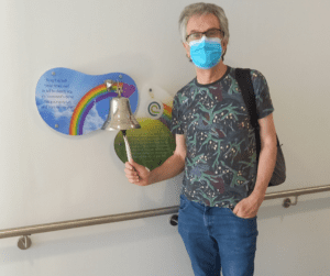 Steve stands in a hospital corridor, ringing a bell. There is a sign behind it with a rainbow. He is wearing jeans and a shirt. He has a mask covering his face.