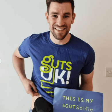 A man with short dark hair and a short dark beard is smiling at the camera. He is wearing a blue, shortsleeved t-shirt and is slightly raising it on one side to show a black stoma bag. The t-shirt has Guts UK's logo on it in green and white. He is holding a sign that says 'This is my #GUTSelfie'