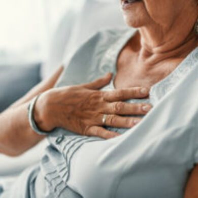 A woman is sat down. She has her hand held up to her chest, indicating pain or discomfort. She is wearing a light blue t-shirt.