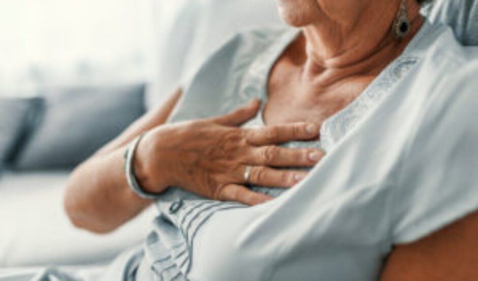 A woman is sat down. She has her hand held up to her chest, indicating pain or discomfort. She is wearing a light blue t-shirt.