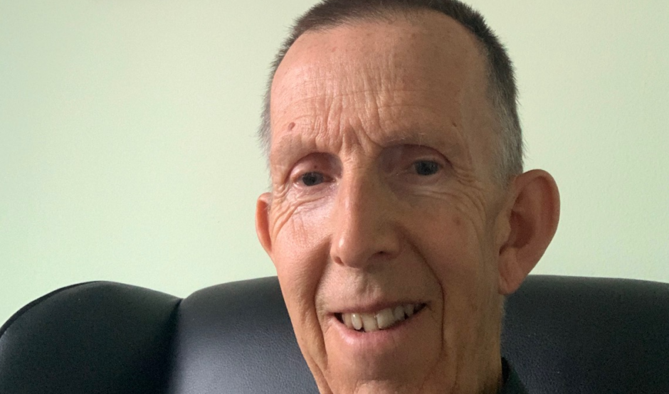 Dave is sat on a black armchair. He is smiling at the camera. He has short dark hair and is wearing a shirt with a collar. The wall behind him is light green and there's a framed picture of a train on the wall behind him.