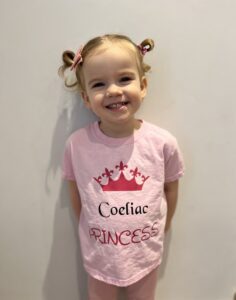 Imogen is smiling at the camera with her ponytails tied into playful loops that stick up in the air. She’s wearing a pink shirt that reads: “Coeliac PRINCESS” with a tiara above the words.