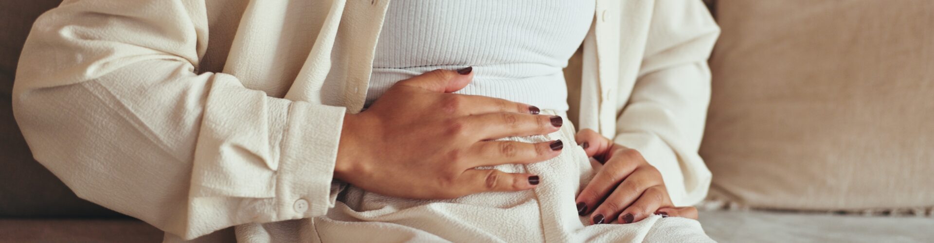 A person wearing beige trousers, a white top and a cream jacket is sat down on a sofa. They are holding their hand over their stomach.