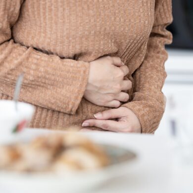 A person's torso is seen, covered by a light brown jumper. They arms are covering their stomach, with their hands gripping their upper abdomen, indicating pain or discomfort.