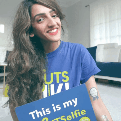 Sharan is smiling at the camera and is wearing a blue Guts UK t-shirt. She holds a board which reads 'This is my #GUTSelfie' and wears #GUTSelfie temporary tattoos on the other arm. She has long, wavy brown hair and wears red lipstick.