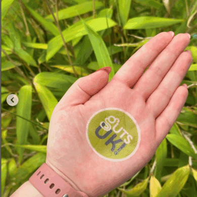A palm of a hand is facing the camera, with a green, circular Guts UK logo on it. There is a pale pink apple watch strap on the wrist and bright green, lush leaves in the background of the photo.