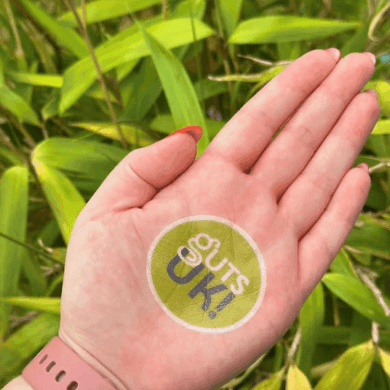 A palm of a hand is facing the camera, with a green, circular Guts UK logo on it. There is a pale pink apple watch strap on the wrist and bright green, lush leaves in the background of the photo.