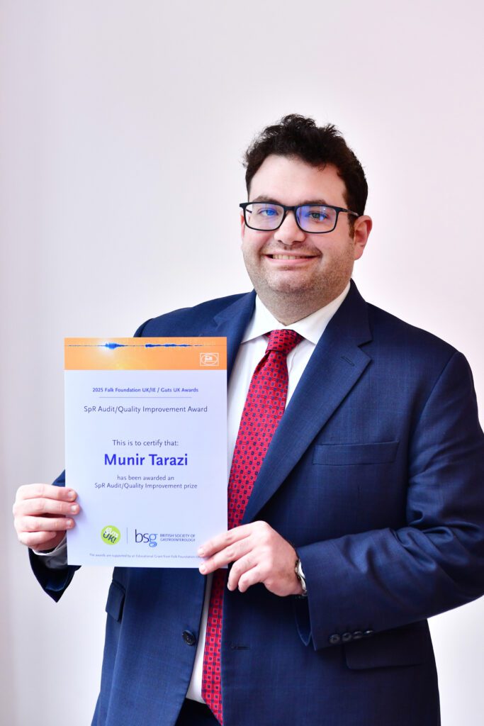 Munir smiles at the camera, holding his certificate. He has short black hair, is wearing black rimmed glasses and wears a navy suit, with a white shirt and red tie underneath.