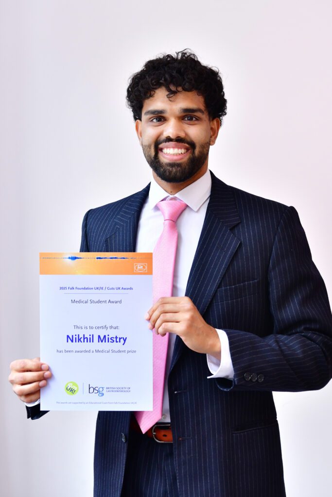 Nikhil smiles at the camera, holding his certificate. He wears a deep navy suit, a white shirt and light pink tie. He has black hair.