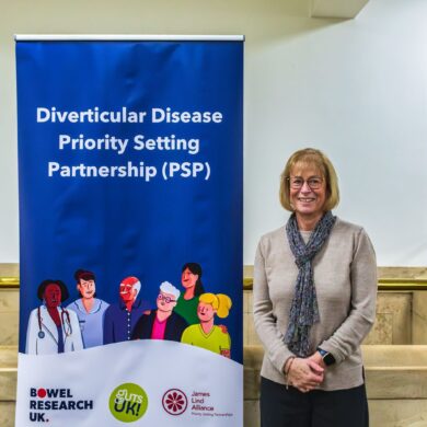 Linda has short, straight, blonde hair styled in a bob, wears black rimmed glasses and wears a beige jumper, blue floral scarf and black trousers. She smiles at the camera, stood next to a narrow roller banner with a Guts UK navy background which has white text on it, reading 