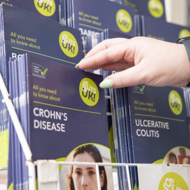 Guts UK's patient information leaflets are displayed in a stand. A hand reaches out to pick up the Crohn's disease leaflet.