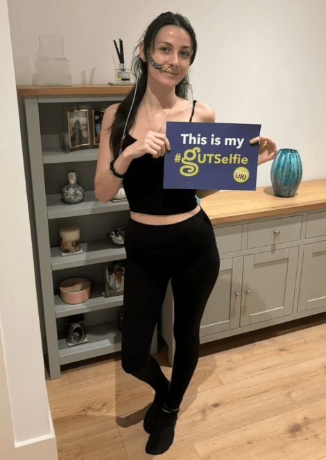 Jessie is wearing a black vest top and leggings, and socks. She has a feeding tube going into her nose, with navy tape holding it to her left cheek. She is holding a #GUTSelfie board and smiles at the camera, in front of a grey bookcase and cabinet.
