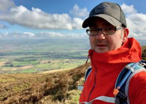 Shawn, wearing glasses, a cap, and a red jacket, stands on a grassy hillside with rolling countryside and a cloudy sky in the background.