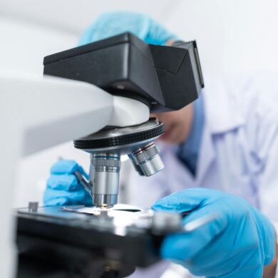 A scientific microscope is in focus along with a researcher's hands, wearing blue, medical gloves. The researcher is blurred but is looking at something on a petri dish under the miroscope, wearing a white lab coat and blue hair net.