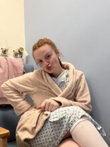 Rachel is wearing a hospital gown and pink robe in a hospital. She is sat in a chair, making a playful face. She has a nasogastric tube and wears compression stockings.