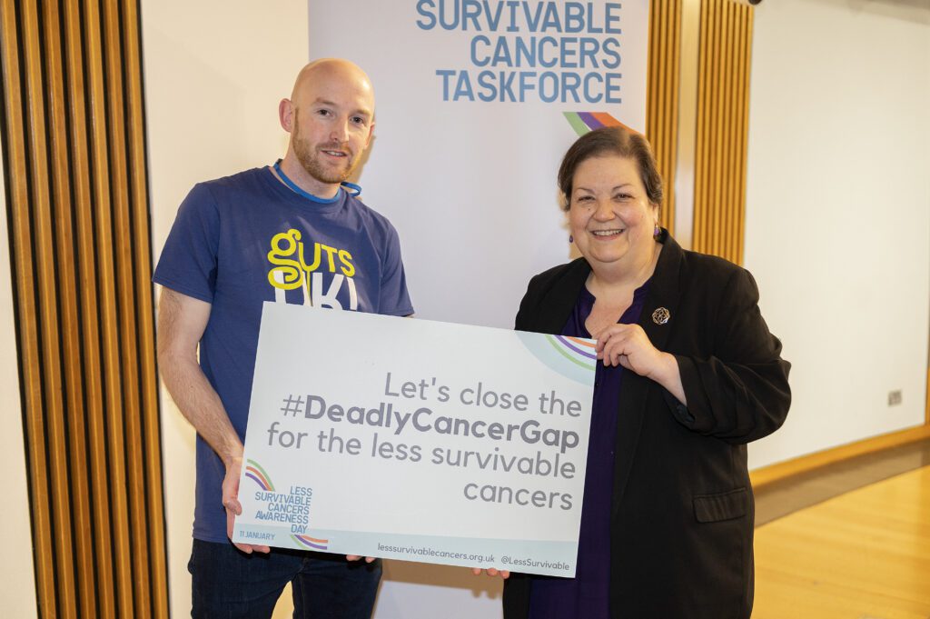 Ross, a family advocate, is pictured with a member of Scottish Parliament, Jackie Baillie, in Scotland. They are holding a Less Survivable Cancers Taskforce sign and smiling at the camera.