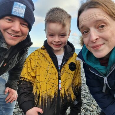 Lisa, Craig and George are smiling at the camera, on a grey, pebbled beach.