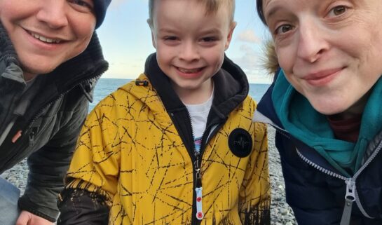 Lisa, Craig and George are smiling at the camera, on a grey, pebbled beach.