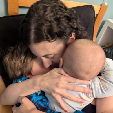 Rachael embraces her two children closely. They are sat on a wooden chair.