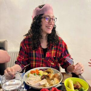 Rachael is wearing a festive pink paper crown and a plaid shirt and in front of a dining table. She is smiling and holding a knife and fork over her plate of Christmas dinner in front of her.