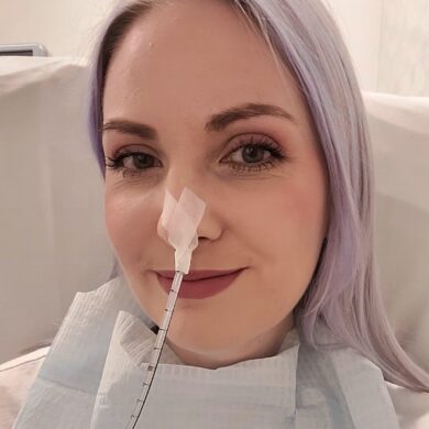 Leanne smiles at the camera in a selfie style photo. She has a feeding tube inserted into her nose and blue roll tucked into the collar of her t-shirt.