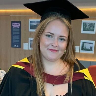 Charlotte wears a long, red patterned skirt, with a black top, and a black graduation gown and cap. Her graduation gown has yellow and red round the shoulders. She smiles, and has long, blonde straight hair.
