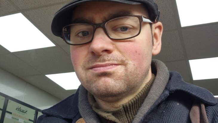 Callan is wearing a navy, thick, blazer jacket and a matching flat cap. He has a pin badge for the poppy appeal on his jacket.