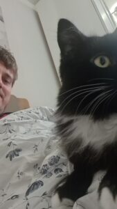 Chris lies under floral bedding, smiling at the camera, while his black-and-white cat, Cola, rests on top of him. Only half of both Chris’s and Cola’s faces are visible, with Cola looking away.