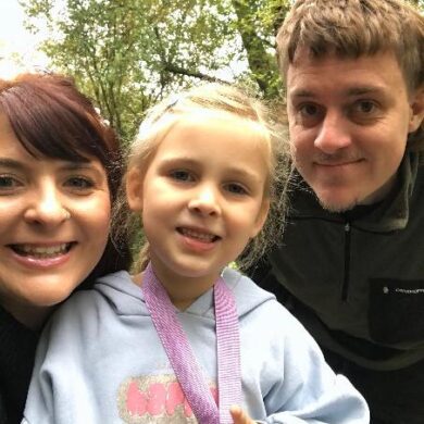 Chris, Rachel, and their daughter Ella-Rose are smiling while standing outdoors in a green, wooded area, looking happy and relaxed.