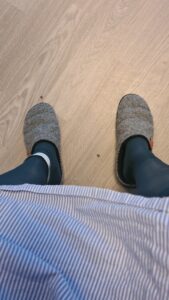 Chris stands on a light wooden floor wearing cozy gray slippers over socks with patterned blue tights. The camera focuses on his feet.