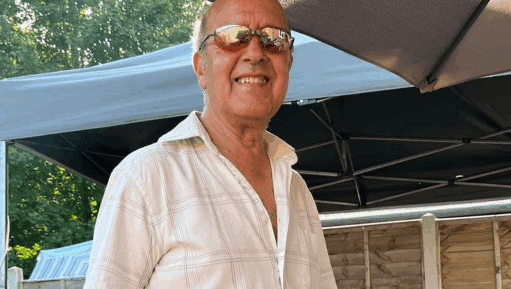 A man in a a white shirt (Charlie) smiles at the camera. Charlie is in the garden and is stood in front of a patio table, he is wearing oven gloves and holding a cooking utensil.