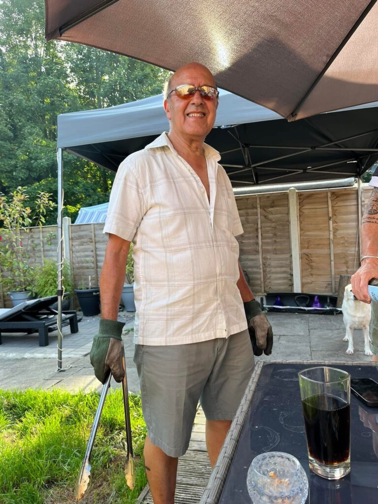 A man in a a white shirt (Charlie) smiles at the camera. Charlie is in the garden and is stood in front of a patio table, he is wearing oven gloves and holding a cooking utensil.
