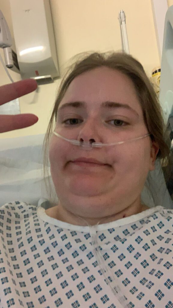 Charlotte takes a selfie in a hospital bed. She holds two fingers up in a "peace" sign and smiles slightly. She has a white hospital gown on with teal diamonds on, an oxygen tube placed on her nose and her blonde hair is up in a ponytail.