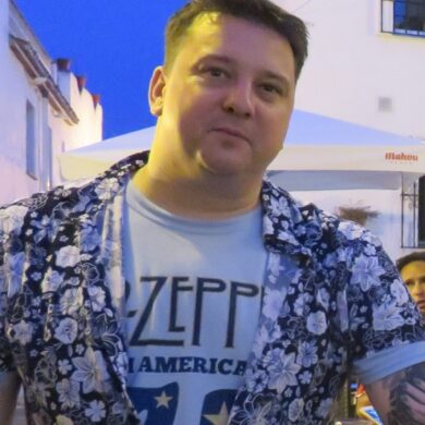 Mark is outside at dusk at wears a white Led Zeppelin t-shirt, with a black and white floral short-sleeved shirt on top, partially buttoned up. He has a sleeve of tattoos on his left arm and short, light brown hair which is spiked up. He smiles slightly at the camera.