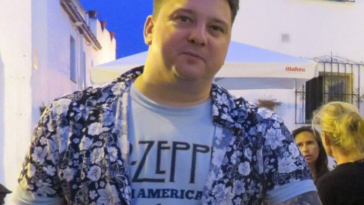 Mark is outside at dusk at wears a white Led Zeppelin t-shirt, with a black and white floral short-sleeved shirt on top, partially buttoned up. He has a sleeve of tattoos on his left arm and short, light brown hair which is spiked up. He smiles slightly at the camera.