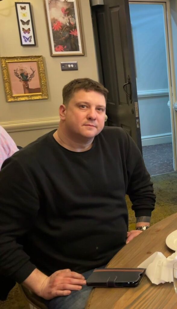 Mark is sat at a table in a restaurant with his mobile phone in front of him. He wears a black jumper and is looking at the camera. His hair is light brown, short and spiky.