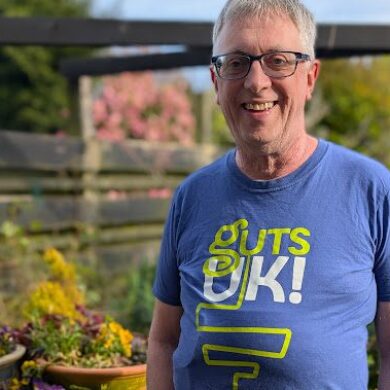 Richard is stood in is garden, wearing a blue Guts UK t-shirt, grey trousers and black rimmed glasses. He has grey hair, and a silver watch on his arm. He has his hands in his pockets and to his left, are a selection of purple flowers, and other shrubs in pots, on a garden table. He smiles at the camera.