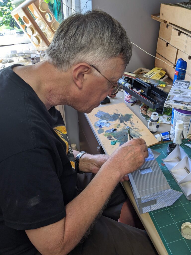 Richard is sat at a table with a craft mat on it. He is putting together a building for his model railway. There is a paint board in front of him, and also various modelling equipment such as paint, pencils and glue. He wears a black t-shirt and also black glasses and has grey hair. The photo is a side profile of him.