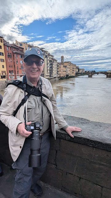 Richard is pictured in Florence, on a bridge, with a river behind him and another bridge in the distant. The sky is bright blue with wispy white clouds and there are yellow and red buildings to his left. He wears a denim cap with pin badges on, black sunglasses, a grey shirt, a light grey jacket, dark grey trousers and is holding his camera which has a long lens on it. He smiles at the camera.