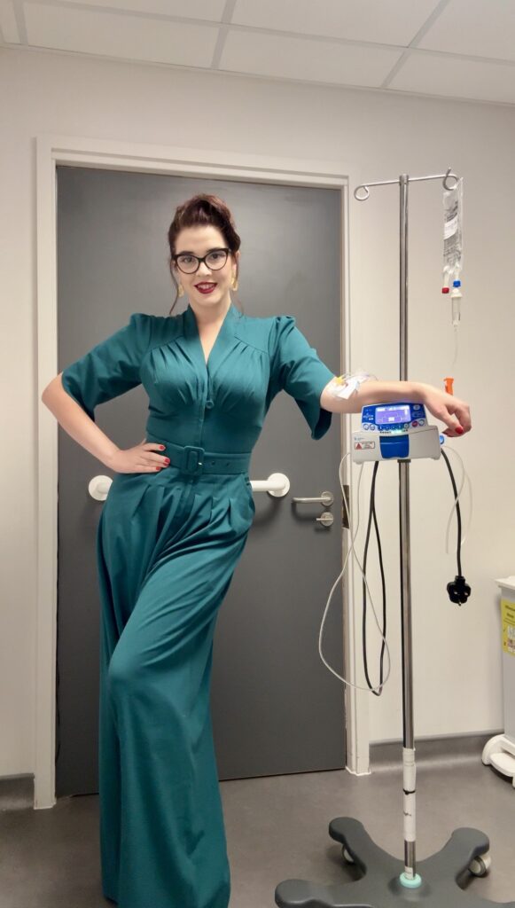 Scarlett is in hospital, receiving treatment for her Crohn's disease via an intravenous drip into her arm. Her drip stand is to the side of her, and she wears a teal vintage, floaty jumpsuit which has three quarter length sleeves. One arm is on her hip and the other rests on top of her treatment machine on her drip stand. She smiles at the camera and has red lips, wears gold hoop earrings and thick, black rimmed oval glasses. Her dark brown hair is up, with a loose strand down the side of her face.