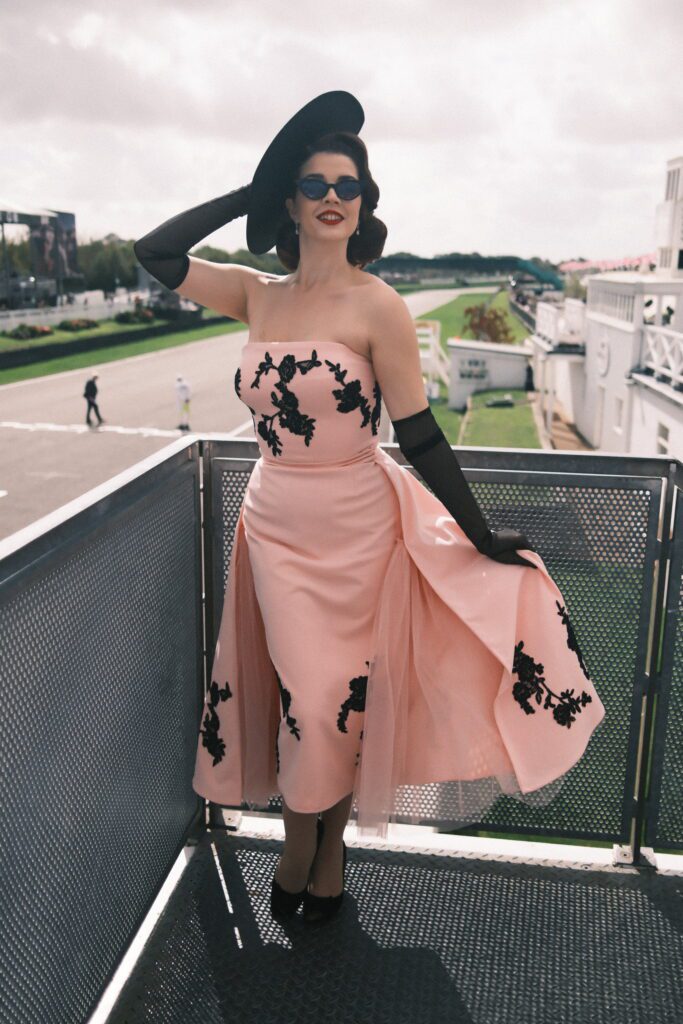 Scarlett is pictured at an outdoor event with a racing track in the background, stood on a balcony. She wears a pale pink, strapless, floaty dress, which goes down to just below her knees. On the dress is black floral detail, and she wears long, black satin gloves, black oval sunglasses, a black hat tilted to one side of her head and black court shoe heels. She has red lipstick on and short dark brown hair, with loose curls, in a bob. She poses proudly.