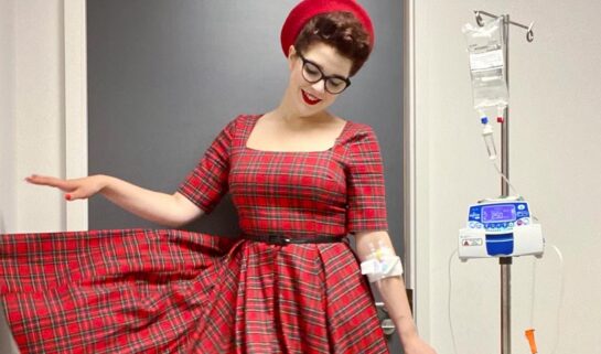 Scarlett is in hospital, connected to an intravenous drip going into her arm, to receive her Crohn's disease treatment. Her drip stand is beside her. She is stood smiling towards the floor, in a three quarter sleeve vintage, red tartan dress and has red nails to match. A thin black belt cinches her dress in at the waist, and she also wears a bright red beret style hat and black patent Mary Jane style heels. She wears red lipstick and thick, black rimmed oval glasses. One arm is down by her side and the other has just let one side of her tartan dress down from her waist, to reveal the full skirt.