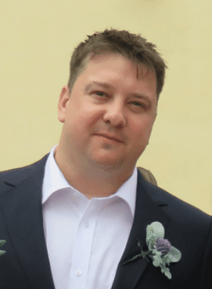 Mark is pictured against a pale yellow background. The photo is cropped from his chest upwards. He wears a black suit jacket, with a light green and purple floral buttonhole piece, and a crisp white shirt underneath. He has short, light brown, spikey hair and smiles at the camera.