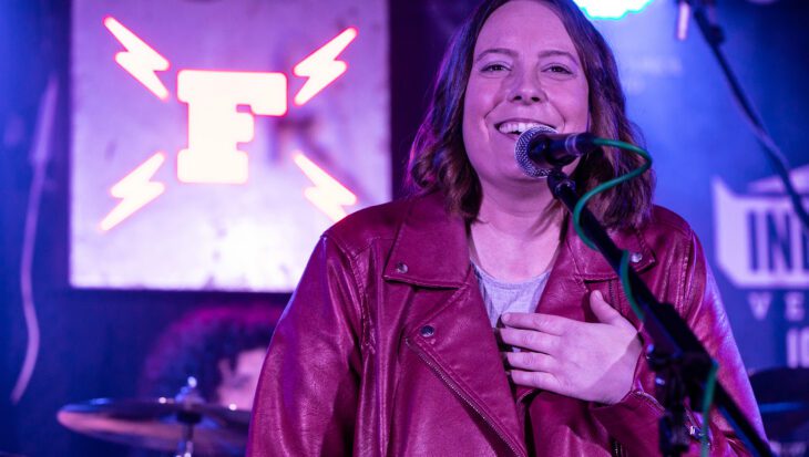 Beth is wearing a burgundy leather jacket and passionately singing into a microphone on stage. She has her left hand on her chest and is standing in front of a sign that says 