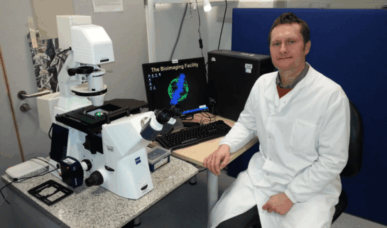 Dr Jason Bruce sits at a desk wearing a white lab coat. He is sat next to science equipment and a computer.