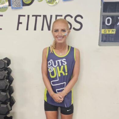 Eryn is standing in a gym wearing a blue Guts UK running vest and black shorts. She also wears black and white trainers.